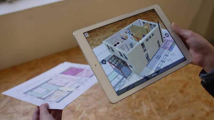 Person using a tablet to view an augmented reality 3D model of a house projected over a printed architectural floor plan.
