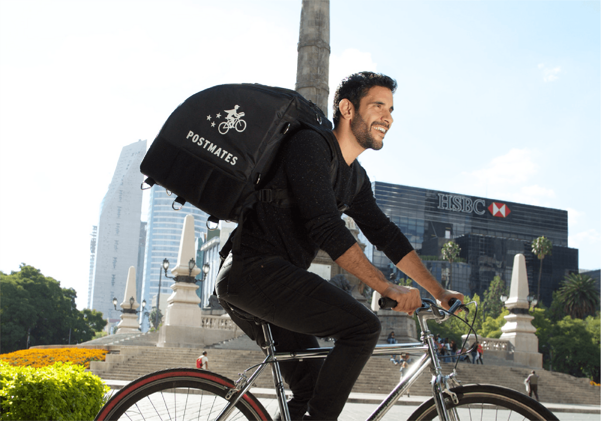 Postmates delivery courier riding a bicycle with a branded delivery backpack in an urban area, passing by a monument and modern buildings.