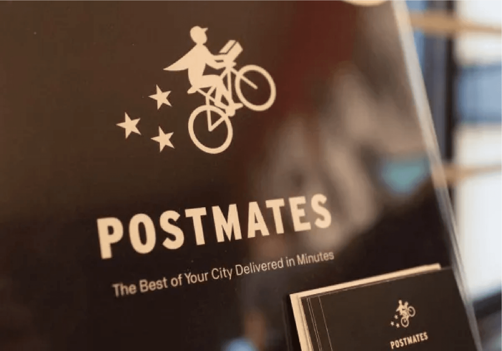 Postmates promotional sign displaying the logo with a delivery cyclist and stars, along with the tagline "The Best of Your City Delivered in Minutes."