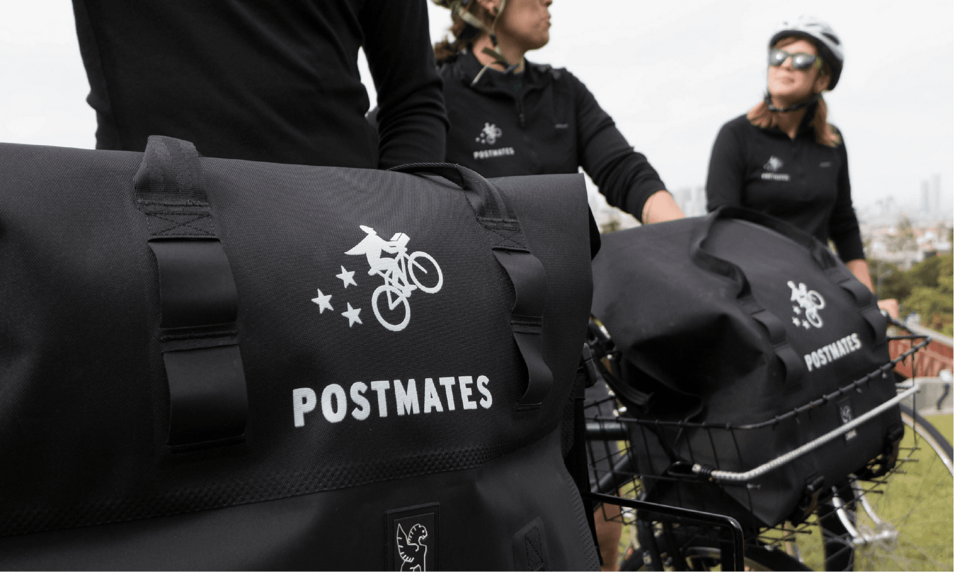 Postmates couriers wearing branded gear and carrying black delivery bags with the Postmates logo, standing with bicycles in an outdoor setting.