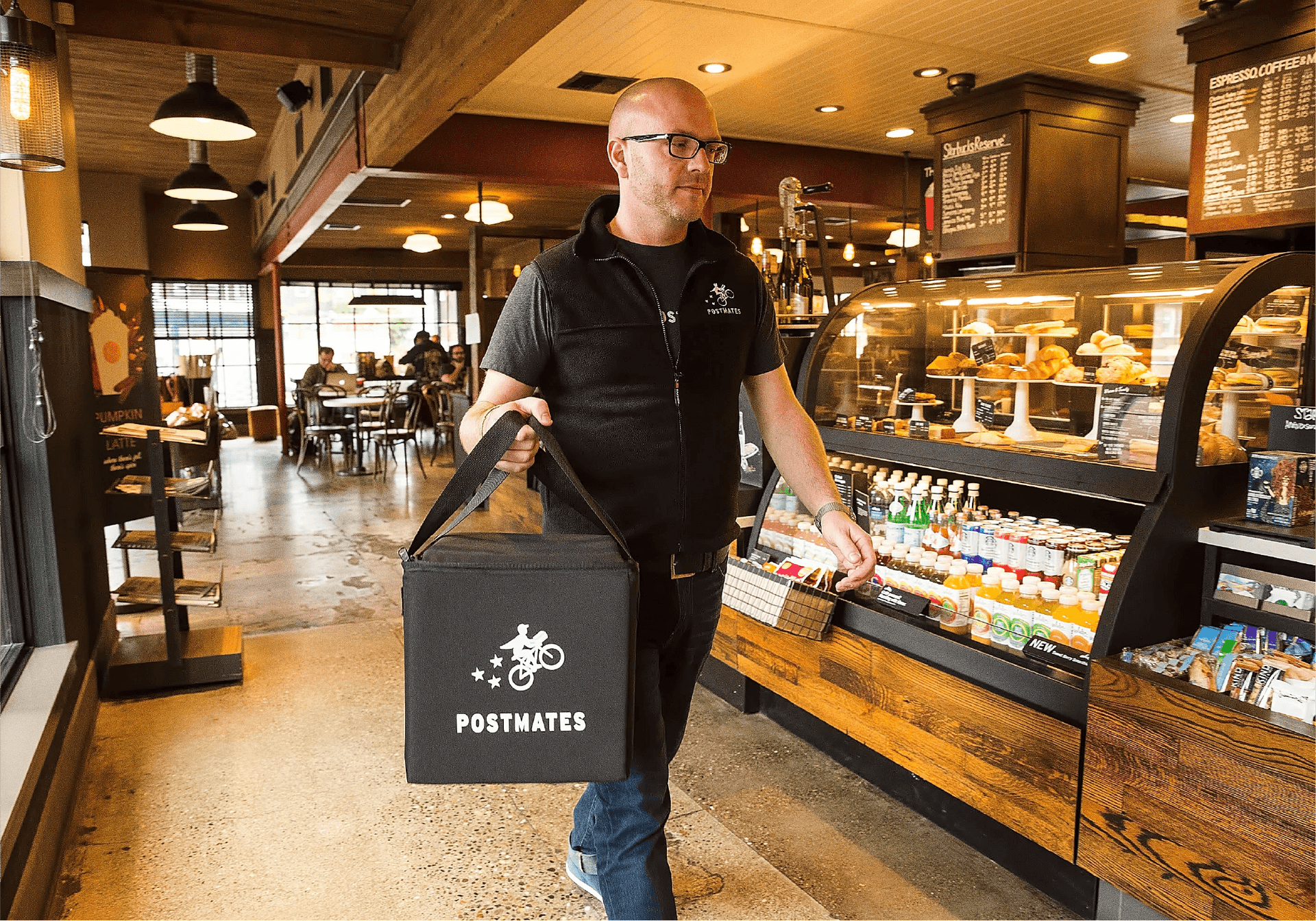 Postmates delivery courier picking up an order inside a café, holding a branded insulated delivery bag, with food and drinks visible in the display case.